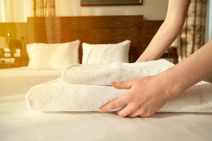 Close-up of hands putting stack of fresh white bath towels on the bed sheet. Room service maid cleaning hotel room. Lens flair in sunlight